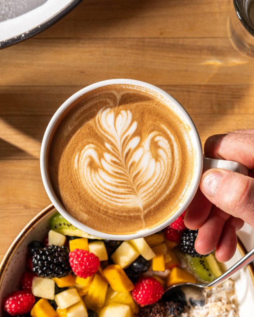Hand holding a latte in a white mug with a bowl of fruit on wooden table
