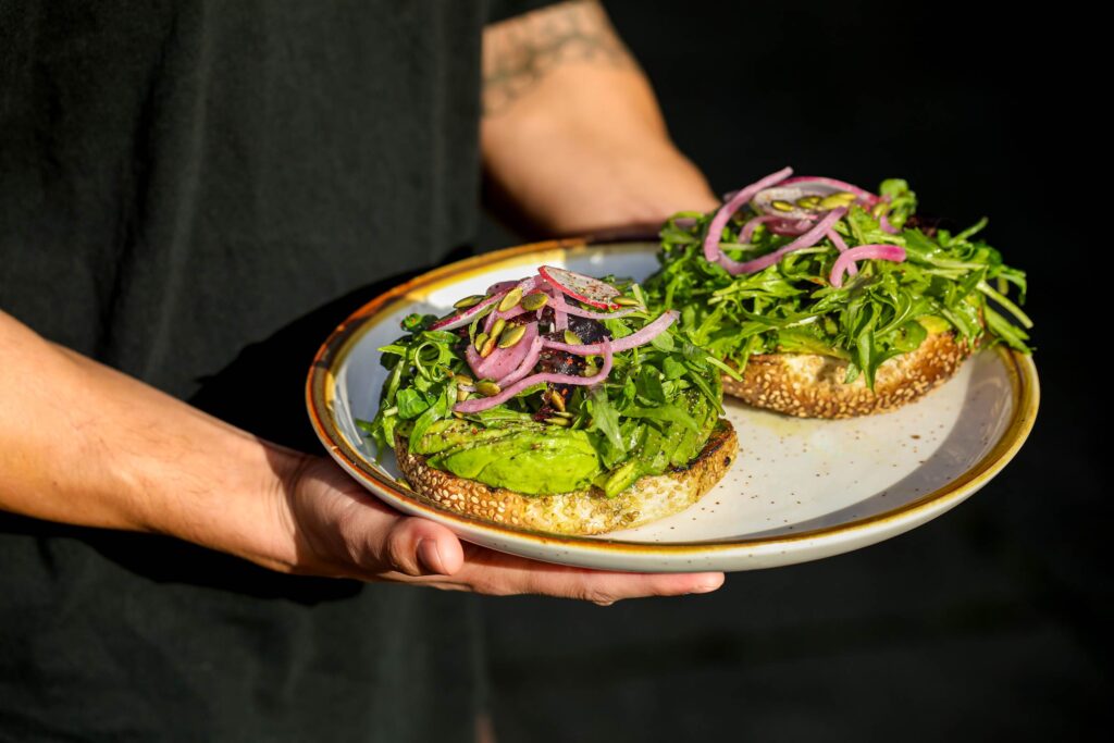 Hands holding plate with open faced sesame bagel topped with avocado, arugula, onions, and seeds
